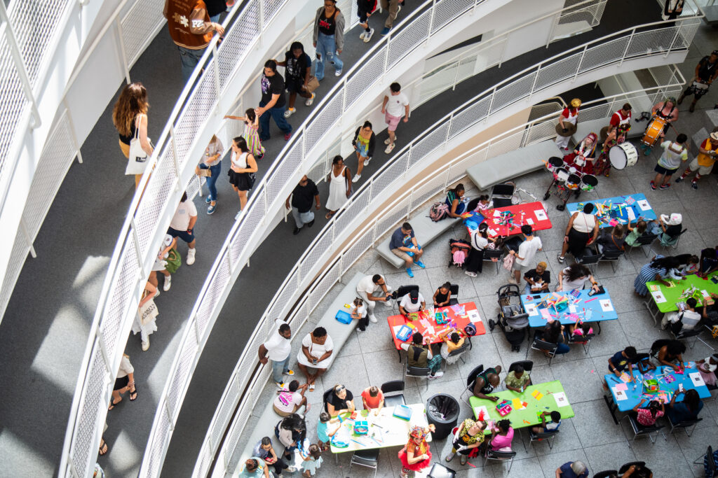A top-down view of a busy, multi-level indoor space. People walk along curved balconies while others sit at colorful tables below, engaged in various activities. The scene is lively and social.