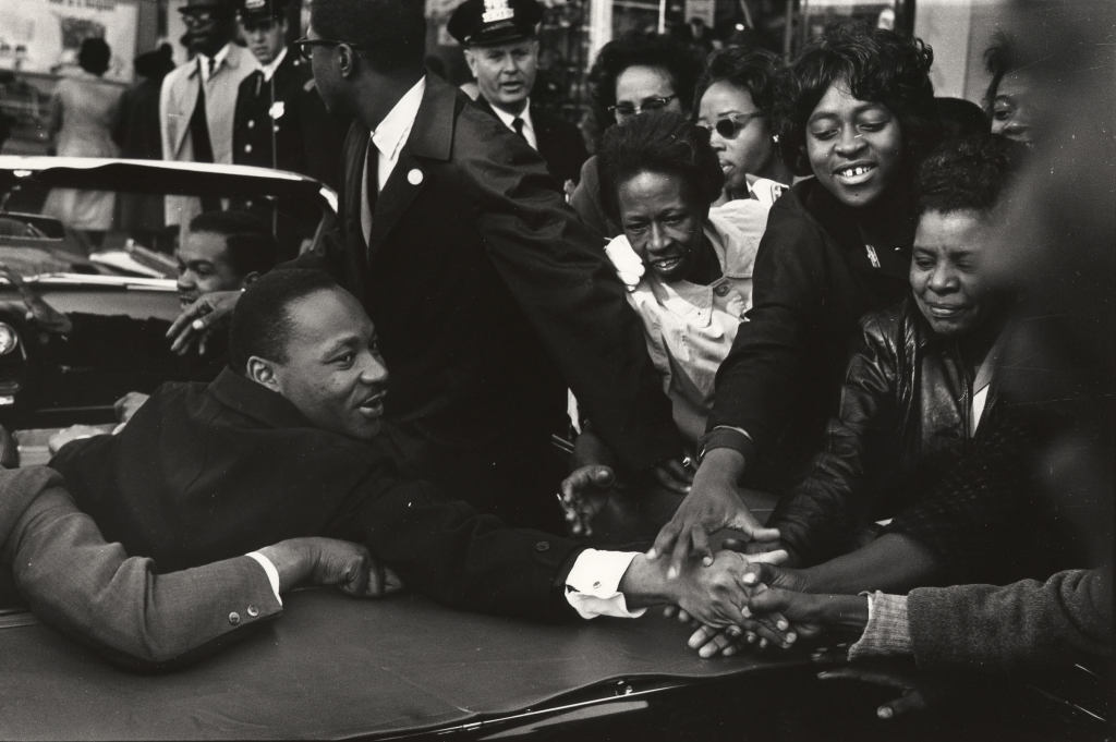 Martin Luther King, Jr. reaches out from the backseat of a convertible to shake hands with smiling protesters in a crowd. White policemen stand in the background.