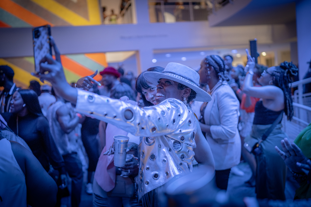 A group of people are at a lively indoor event. In the foreground, two women are taking a selfie, one wearing a silver jacket and wide-brim hat. The background features a vibrant wall with diagonal stripes and other attendees are engaged in conversation and taking photos.